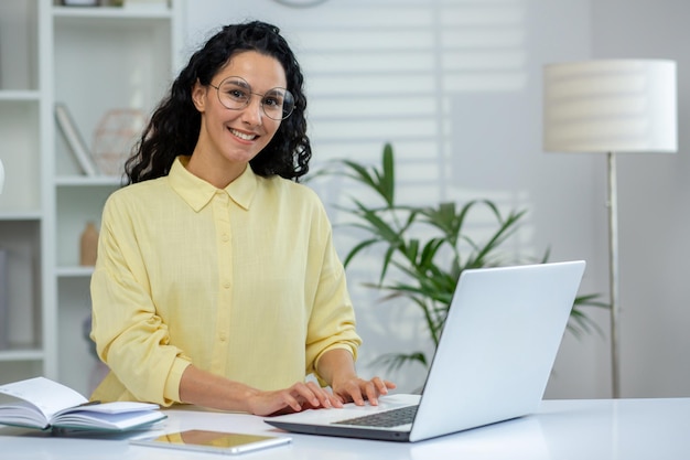 Smiling woman in glasses working on a laptop in a bright modern office with plants in the background