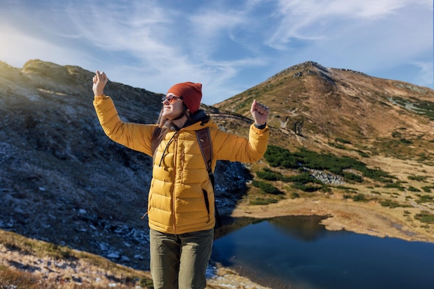 Smiling woman hiker in yellow down jacket with backpack is hiking mountains at sunny autumn day