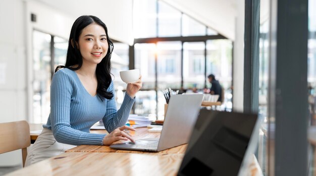 Smiling woman holding coffee cup and using laptop while sitting at coworkspace