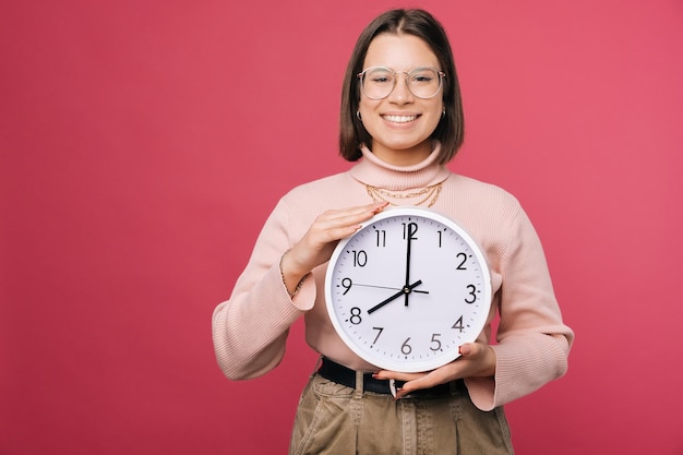 Smiling woman is holding a white round clock carefully over pink backdrop