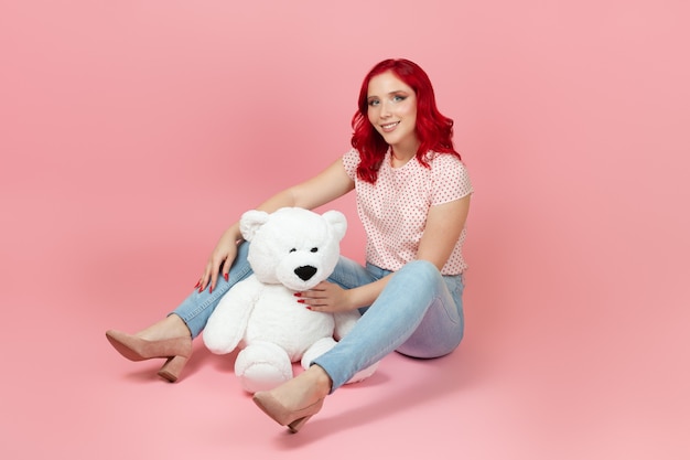 Smiling woman in jeans with red hair with a large white teddy bear sitting on the floor