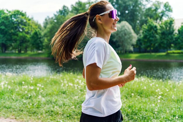 Photo smiling woman jogging in a lush park by a serene lake on a bright day