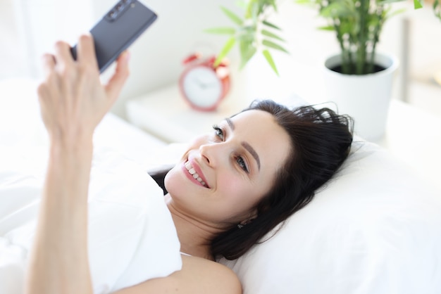 Smiling woman lies in bed and looks into smartphone