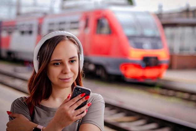 Smiling woman listens to music with headphones using smartphone on railway platform