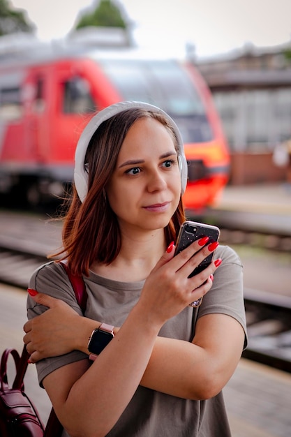 Smiling woman listens to music with headphones using smartphone on railway platform