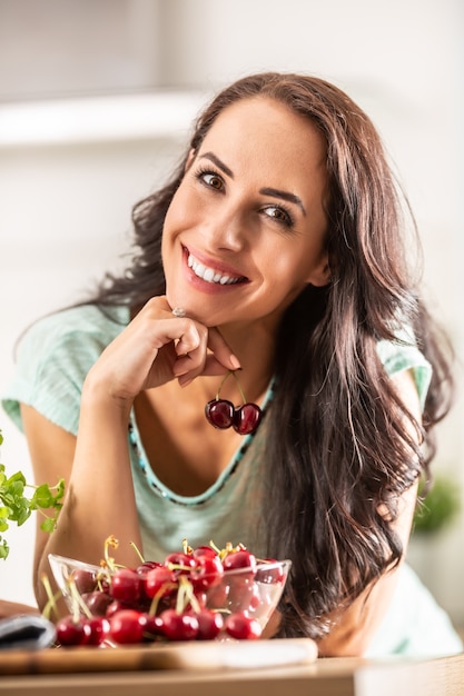 Smiling woman picks two cherries from a seethrough bowl indoors.