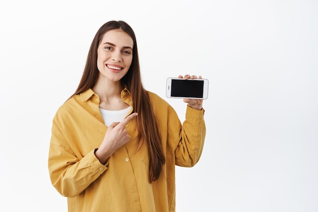 Smiling woman pointing at smartphone showing application online shop website or video game advertisement on screen standing over white background