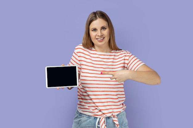 Smiling woman pointing at tablet empty screen for adv looking at camera with toothy smile