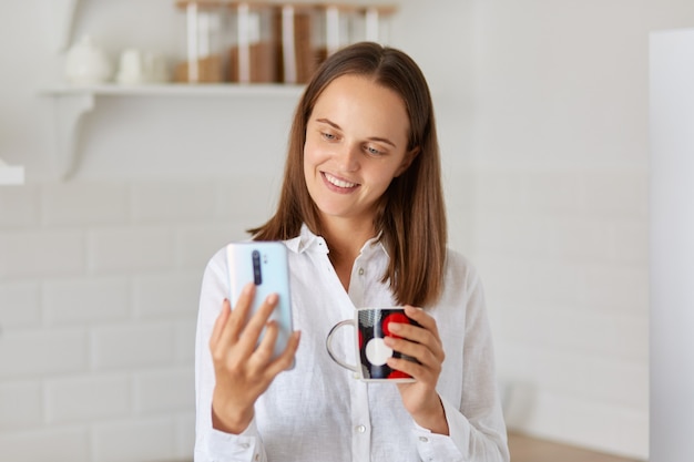 Smiling woman posing in light kitchen and using smart phone while drinking coffee or tea in the morning, looking at cell phone display, having video call or taking selfie.