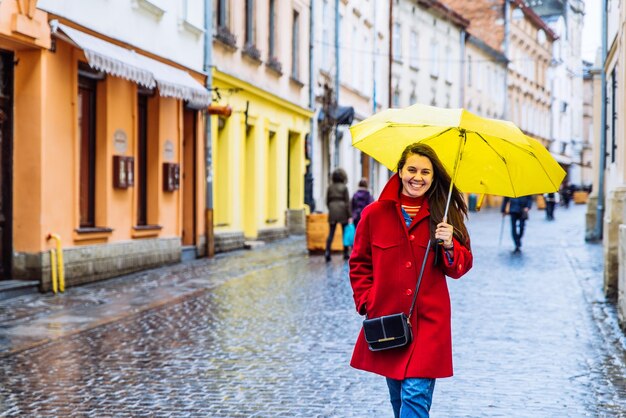 Smiling woman in red coat with yellow umbrella walk by city street