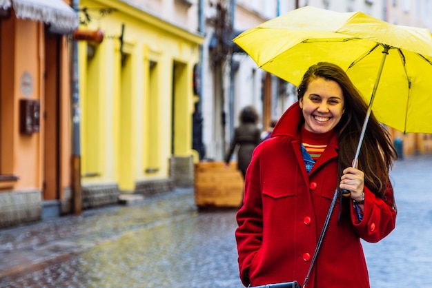 Smiling woman in red coat with yellow umbrella walk by city street