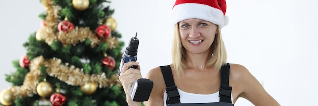 Smiling woman repairman in uniform and santa claus hat holding drill in hands near christmas