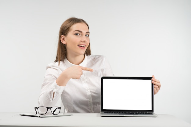 Smiling woman sits at the table and point to the screen  looking at the camera