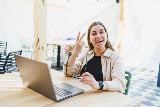 Smiling woman sitting at outdoor cafe table with laptop talking on phone and cup of coffee smiling woman in glasses enjoying telecommuting in cafe or studying online