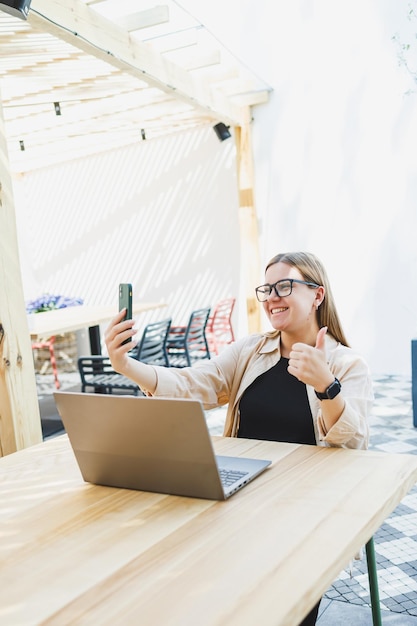 Smiling woman sitting at outdoor cafe table with laptop talking on phone and cup of coffee smiling woman in glasses enjoying telecommuting in cafe or studying online