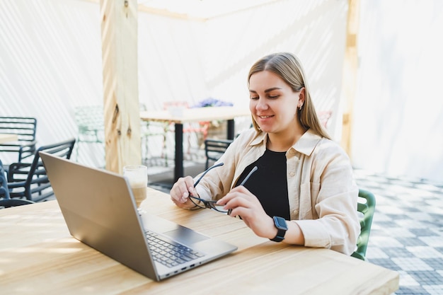 Smiling woman sitting at outdoor cafe table with laptop talking on phone and cup of coffee smiling woman in glasses enjoying telecommuting in cafe or studying online