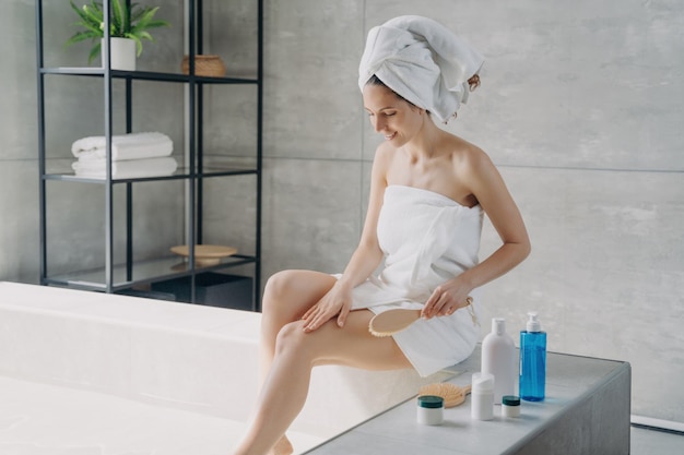 Smiling woman in spa setting using a body brush for skin health wrapped in towels with care products on display