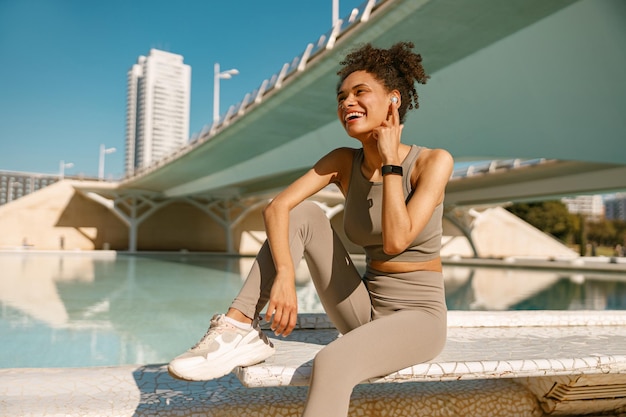 Photo smiling woman in sportswear have a rest after workout outside sitting on bridge background