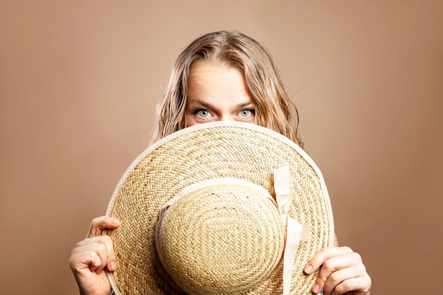 Smiling woman in a summer romantic dress covers her face with a beach straw hat Vacation and tourism Beige background Closeup