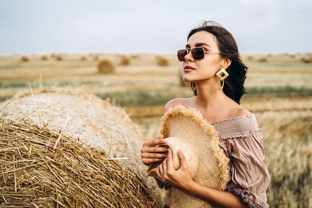 Smiling woman in sunglasses with bare shoulders on wheat field and bales of hay