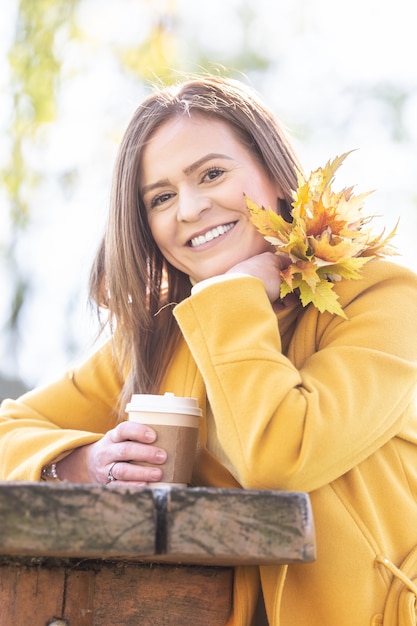 Photo smiling woman on a sunny autumn day outside holds a cup of coffee and couple of yellow leaves.