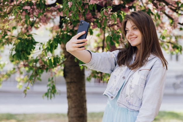 smiling woman taking selfie self portrait photos on smartphone model posing on park sakura trees