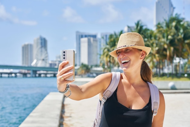 Smiling woman taking selfie with smartphone in sunny urban backdrop palm trees and cityscape of Miami