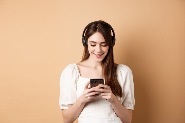 Smiling woman texting message on phone and listening music in wireless headphones, watching video on smartphone, beige background.