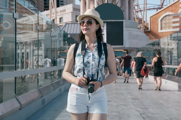 smiling woman traveler in hat holding vintage camera in las vegas modern city usa. Hipster girl tourist photographer walking outdoors on pedestrian overpass. Travel summer time vacation concept.