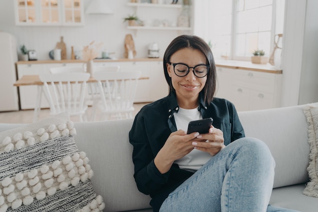 Smiling woman uses smartphone chatting in social networks shopping online sitting on couch at home