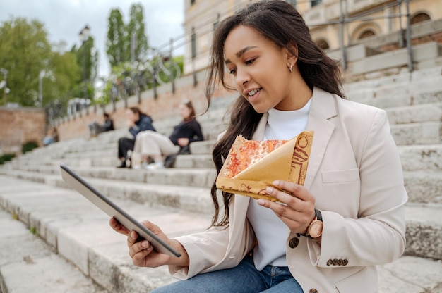 Smiling  woman using digital tablet looking at screen, holding slice of tasty pizza outdoors