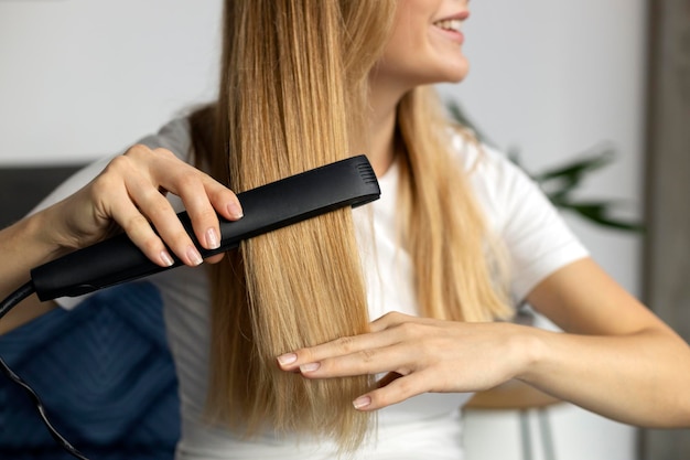 Smiling woman using hair straightener at home, selective focus on hair. Beauty, hair care concept