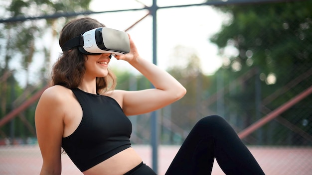 Smiling woman using VR headset in a tracksuit while sitting on yoga mat on a sports field in a park