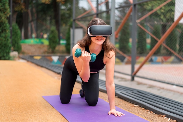 Photo smiling woman in vr headset in a tracksuit doing exercises with dumbbells on a yoga mat on sports