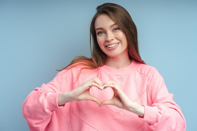 smiling woman wearing pink sweatshirt with toothy smile gesturing heart with fingers