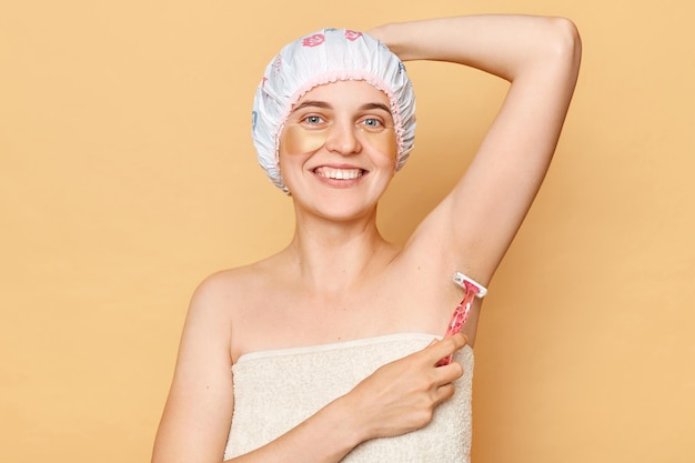 Smiling woman wearing shower cap standing isolated over beige background doing depilation with razortaking care of her armpit