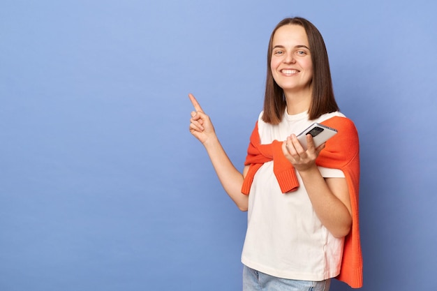 Smiling woman wearing white tshirt and jumper over neck standing isolated over blue background holding smart phone pointing at advertisement area