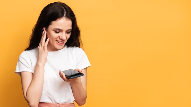 smiling woman in a white top holds a smartphone on orange background with copy space