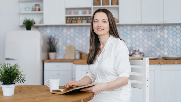 Smiling woman with a book and a cup of coffee sitting in her kitchen