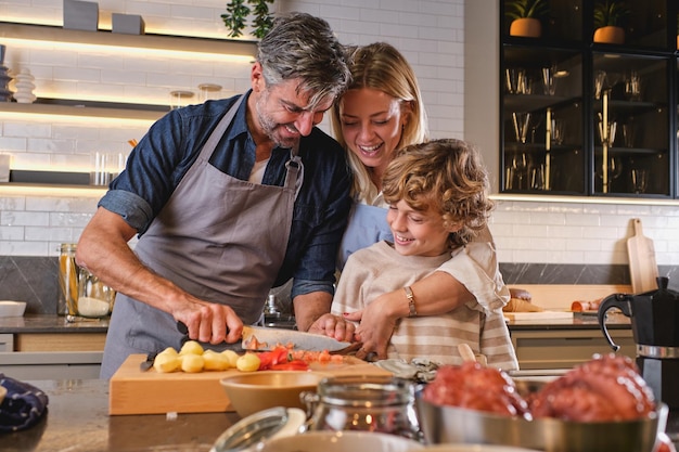 Smiling woman with cute little son standing near kitchen counter and watching father chopping tomato