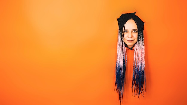 Smiling woman with dreadlocks looking at camera Cheerful young female with multicolored hair looking at camera in the hole on orange background in studio