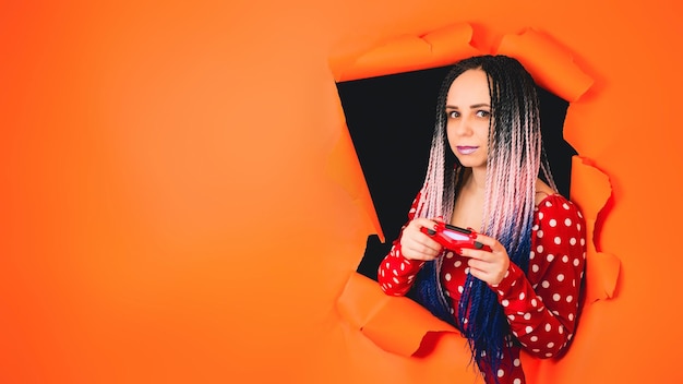 Smiling woman with gamepad and playing video game A stylish girl gamer in a bright red dress playing computer games through a hole in the orange wall in the studio