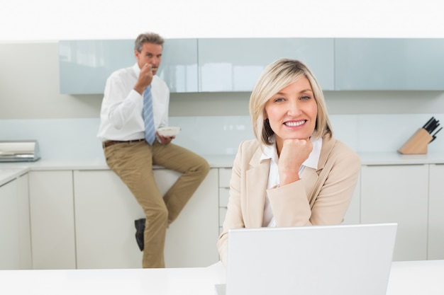 Smiling woman with laptop and man in background at kitchen