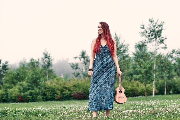 Smiling woman with red hair in a blue dress stands in the park on a summer day. Image in copy space.