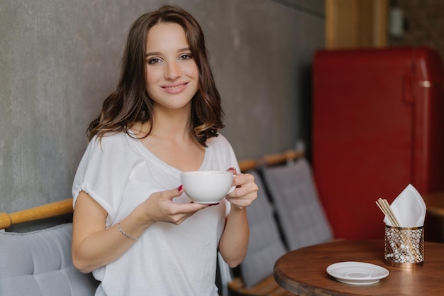 Smiling woman with tender smile holds cup of tea or coffee enjoys good rest poses in cozy cafe or restaurant waits for friend dressed in casual outfit People rest and lifestyle concept