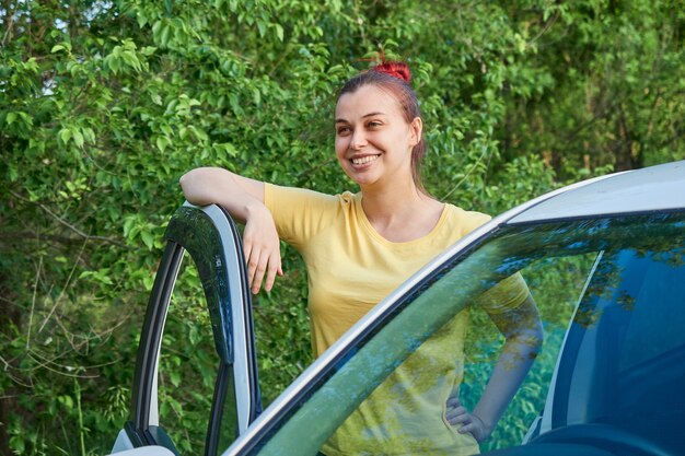 Smiling woman in yellow t-shirt stands near her car on the side of the road opening the door.