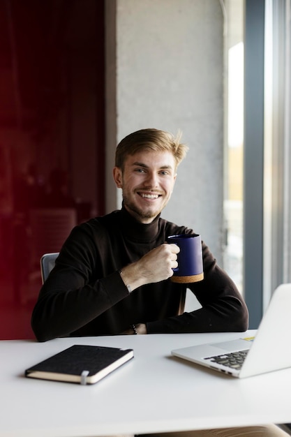 Smiling worker holding cup with drink sitting at workplace looking at camera