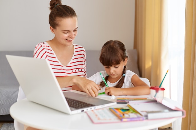 Smiling young adult woman wearing striped casual clothing helping her little daughter to do homework, distance education during quarantine, online lesson.