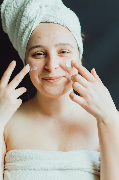 Smiling young adult woman with towel using facial cream after having a shower Skincare wellness of happy face skin glow and sunscreen Daily routine to protect delicate skin