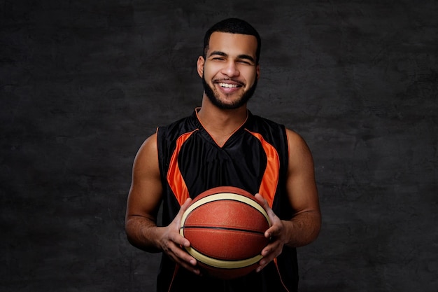 Smiling young African-American basketball player in sportswear isolated over dark background.